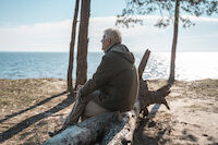 Senior man sitting at the tree log at the seashore and looking at the water surface