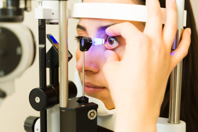 Eine Frau sitzt an der Spaltlampe. Der Untersucher hält mit den Fingern ihr Auge offen, das Messköpfchen des Goldmann-Tonometers nähert sich dem Auge